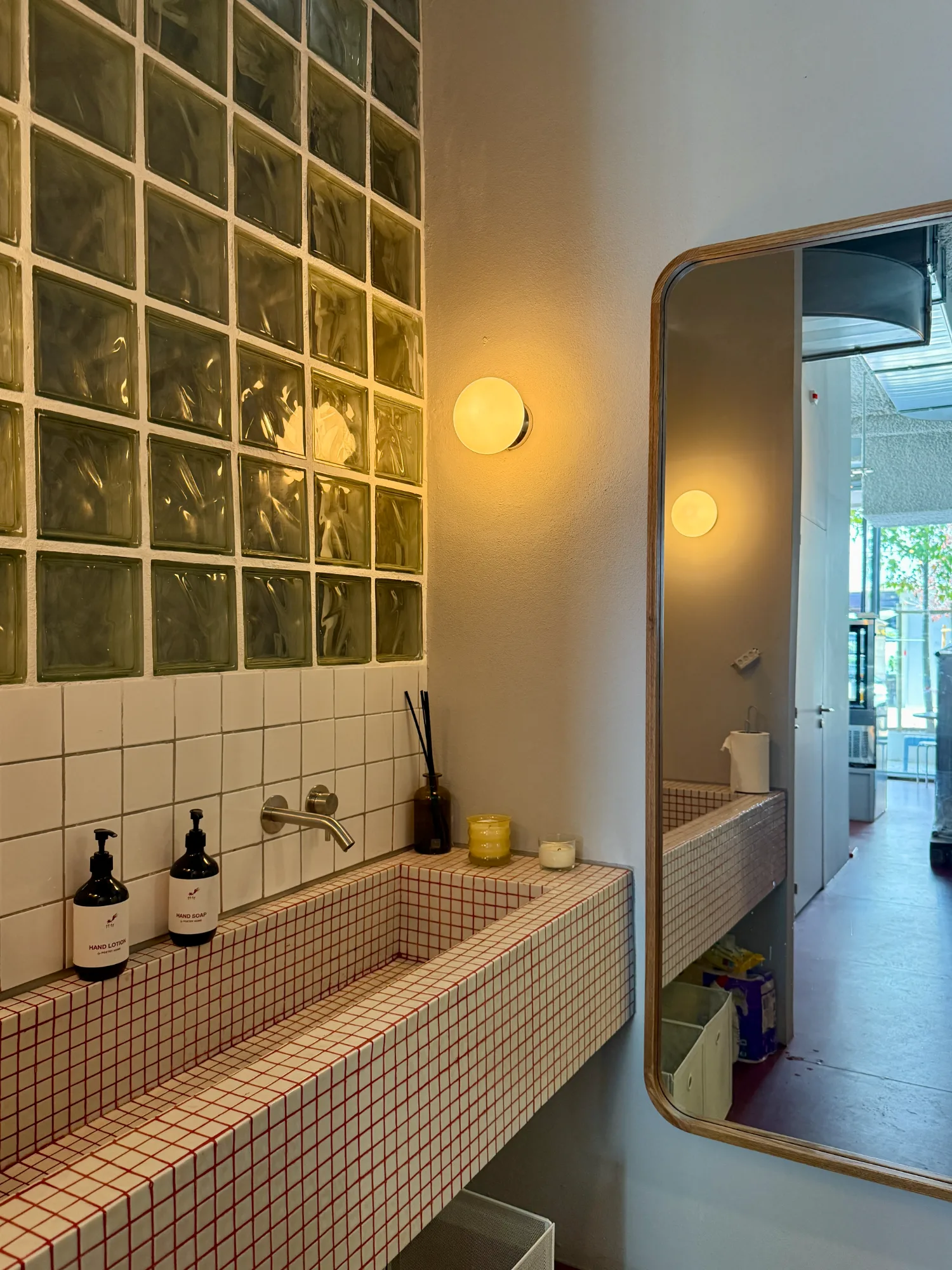 Cafe restroom with custom tiled trough sink, red grout, wall sconce, and glass block partition