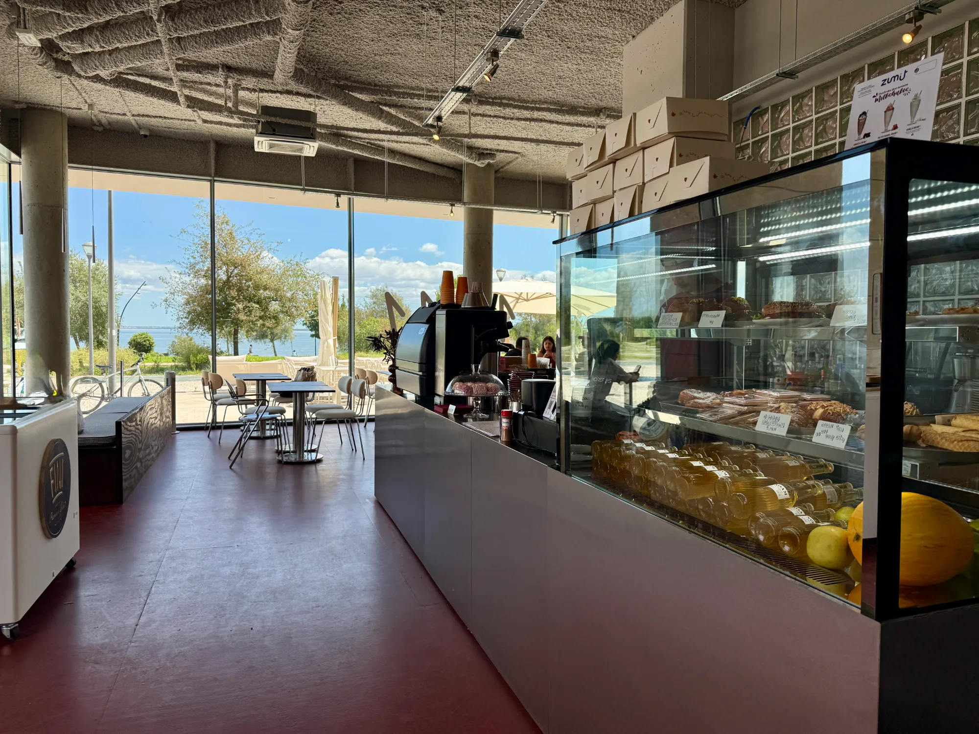 Cafe counter with glass pastry display case and panoramic windows overlooking sunny outdoor promenade
