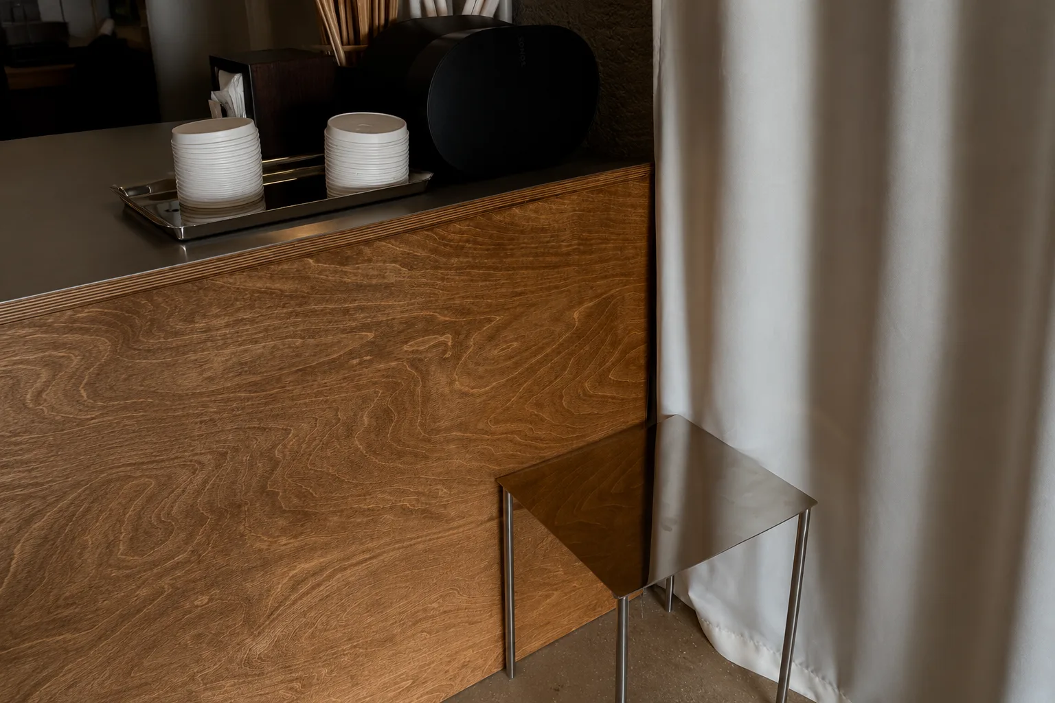 Detail of wooden service counter with stainless steel top, small metal stool, and white curtain