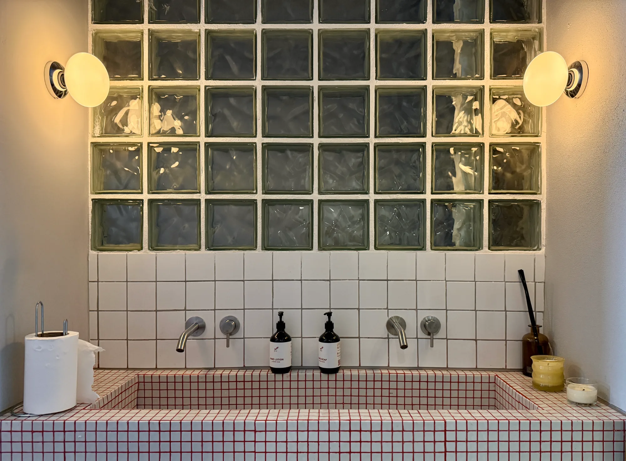 Symmetrical view of cafe washroom with grid-tiled trough basin, double faucets, and glass block wall